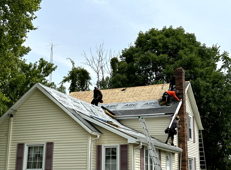 workers replacing the roofing of a beige house with trees around